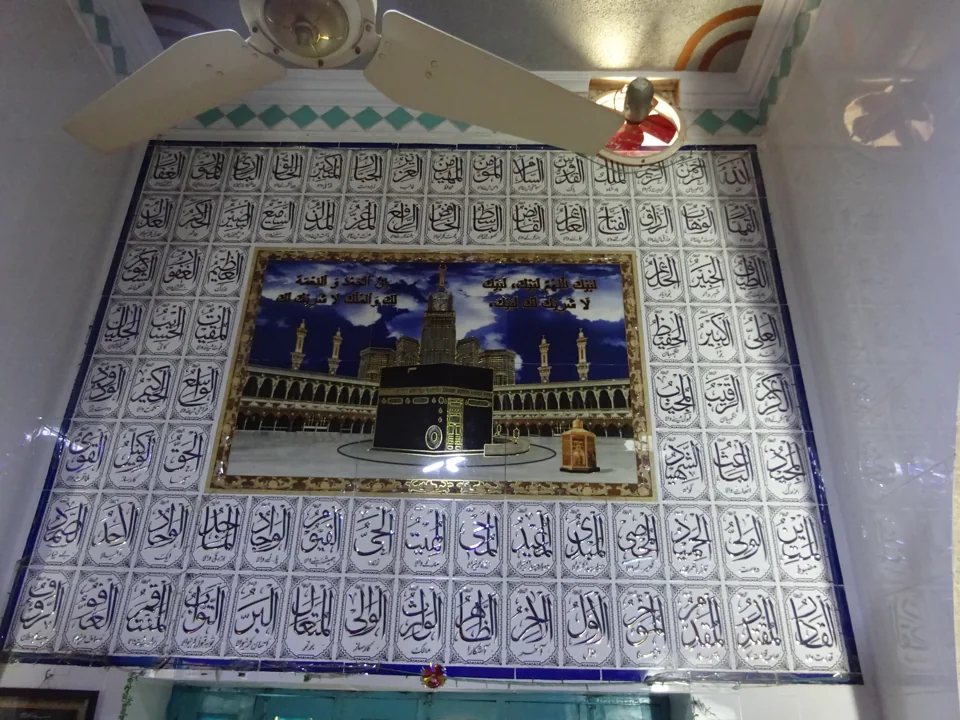 A tiled interior wall with Arabic calligraphy and an image of the Kaaba, beneath a ceiling fan.