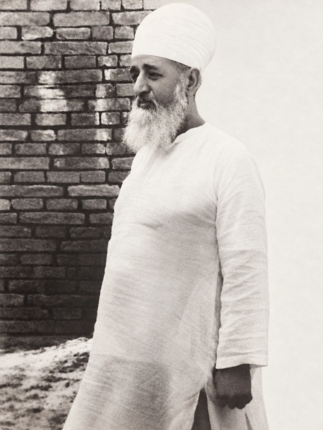 Khwaja Fazal Karim Sahib (RA) in a white turban and traditional dress, standing beside a brick wall.